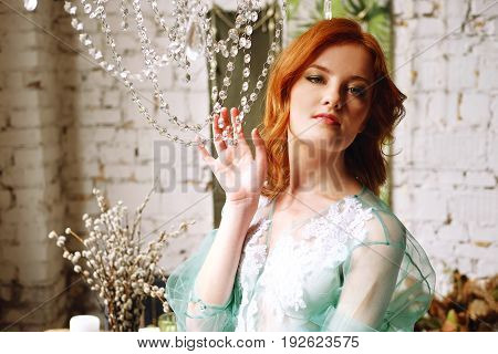 Tender redhead woman in transparent green peignoir and white lingerie on the floor near her bed in bedroom waiting for a man