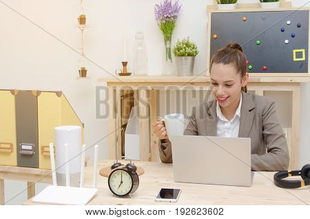 asian young business woman beautiful smile working on laptop notebook computer on wood table with alarm clock headphone smartphone wireless router document folder coffee cupsunlight effect