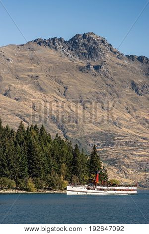 Queenstown New Zealand - March 15 2017: historic white steamboat Earnslaw sails on dark blue Wakatipu Lake under blue sky. Cecil peak brown mountain range and part forest in back.