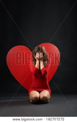 Vertical shot of a heartbroken woman crying sitting on the floor near big red heart symbol symbolic sign breakup relationships sadness depression depressed unloved pity unhappy emotions concept.