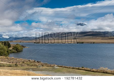 Middle Earth New Zealand - March 14 2017: Wide blue Lake Clearwater in front. Volcano Potts peaking through the white clouds in a blue sky in the back. Green and beige vegetation.