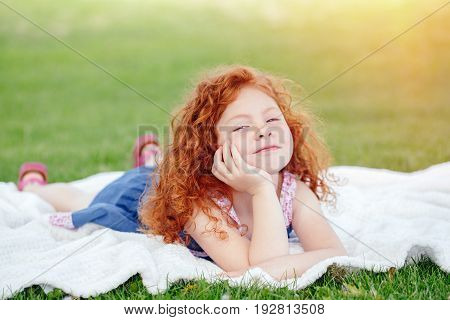 Portrait of cute funny happy smiling little red-haired Caucasian girl child in blue dress lying on green grass in park outside dreaming thinking lifestyle childhood concept