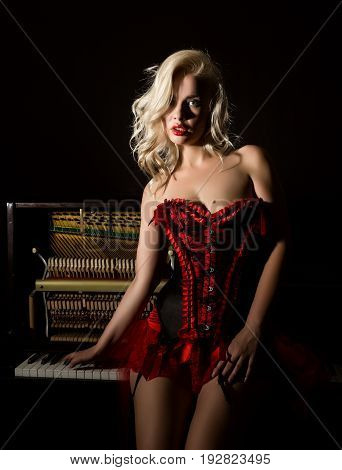 beautiful blonde musician in red corset standing near the piano. Vintage style beautiful woman.. Old fashioned makeup and retro finger wave hairstyle.