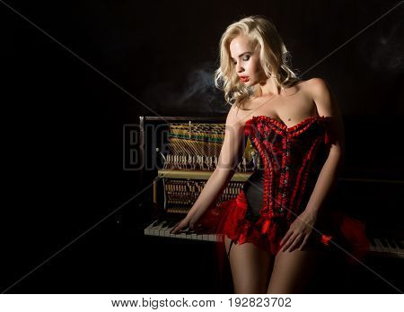 beautiful blonde musician in red corset standing near the piano. Vintage style beautiful woman.. Old fashioned makeup and retro finger wave hairstyle.
