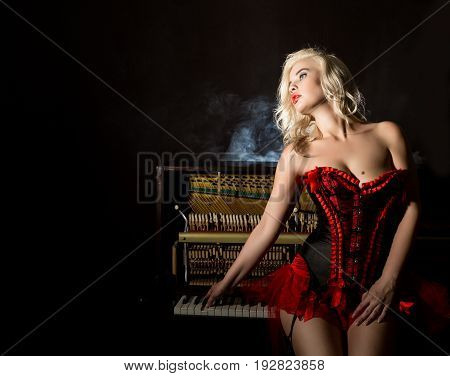 beautiful blonde musician in red corset standing near the piano. Vintage style beautiful woman.. Old fashioned makeup and retro finger wave hairstyle.