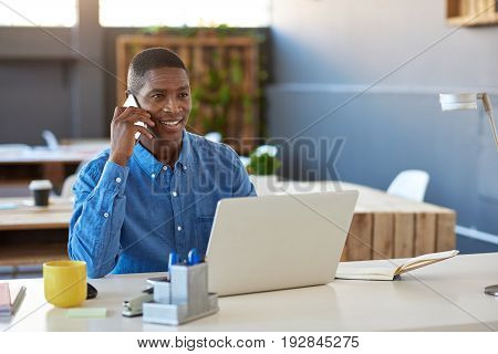 Smiling young African businessman sitting confidently at his desk in a large modern office working on a laptop and talking on a cellphone