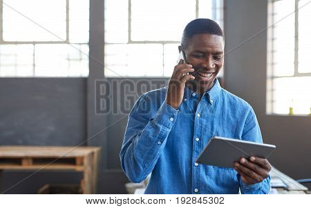 Smiling young African businessman standing in a large modern office working on a digital tablet and talking on a cellphone