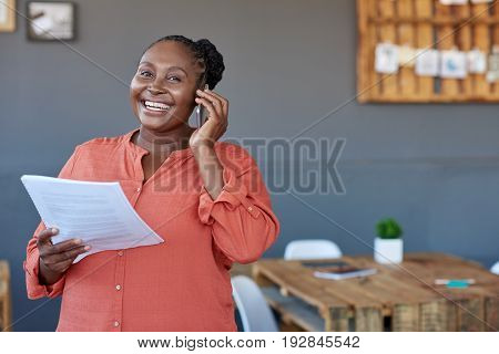 Portrait of a casually dressed young African businesswoman reading paperwork and talking on a cellphone while standing in large a modern office