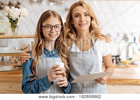 Happy family. Beautiful cheerful mother and daughter standing near the counter of their own coffee bar and posing for the camera with the girl holding a coffee cup and the woman hugging her