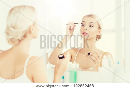 beauty, make up, cosmetics, morning and people concept - young woman applying eyeshade with makeup brush and looking to mirror at home bathroom