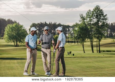 Multiethnic Group Of Golfers Holding Clubs And Talking While Standing On Green Grass