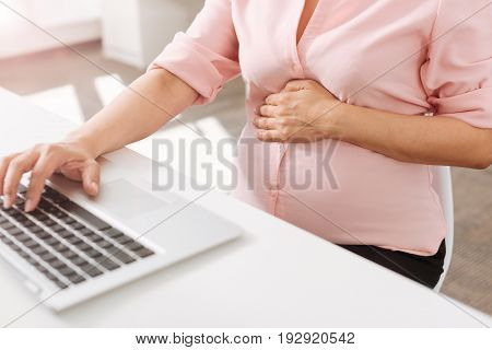 Take care about your future. Pleasant pregnant woman sitting at the table and working in the office while using her laptop
