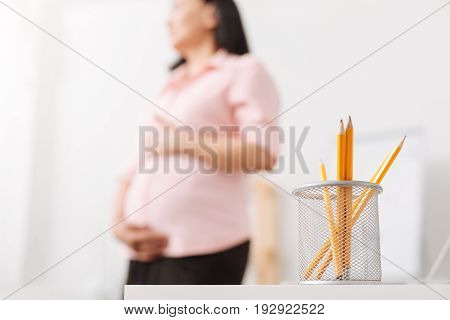 Office stationery. Selective focus of pencils on the office table while pregnant woman standing in the background and touching her belly