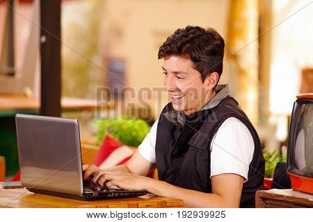 Smiling happy handsome young man at the bar working in his computer on a wooden table.