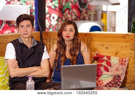 Young couple at the bar spending time together looking at camera, with his computer on a wooden table.