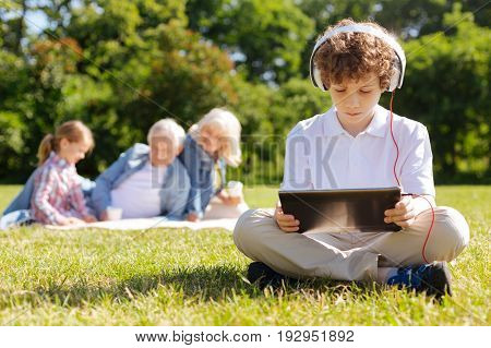 Serious mood. Handsome teenager wearing headphones and crossing legs while sitting on the grass