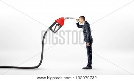 A businessman on white background staring closely at a large red gas nozzle attached to a black hose. Gas station business. Delivery expenses. Fuel market.