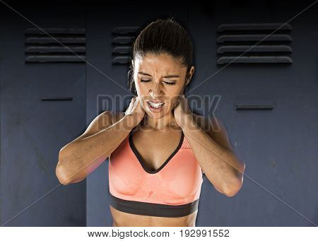 young attractive hispanic fitness woman touching and grabbing her neck and upper back suffering cervical pain isolated on gym locker room background in sport injury and body health care concept