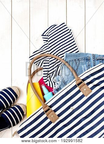 Beach women's clothes and a bag on a white background. Denim shorts summer shoes Striped T-shirt Sunscreen Top view White background