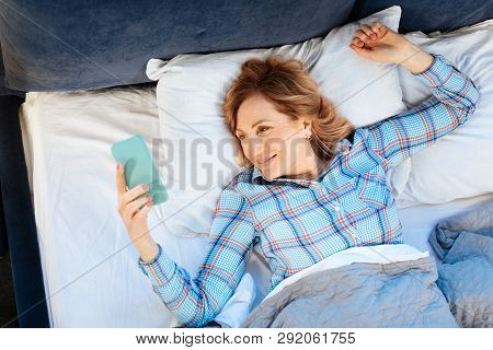 Smiling Positive Adult Lady Lying On Her Back And Holdying Mobile Phone