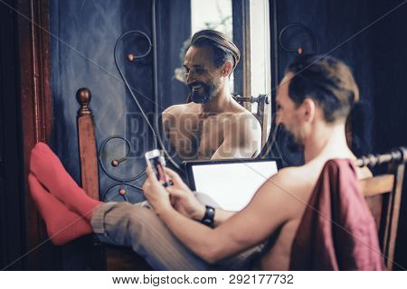 A Handsome Man With A Beard Is Sitting At His Desk At Home. Throwing His Legs On The Table And Leafi