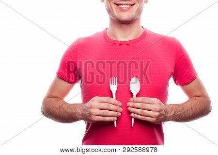 Smiling Handsome Guy Holding Plastic Fork And Spoon Near His Stomach Over White Background. Studio S