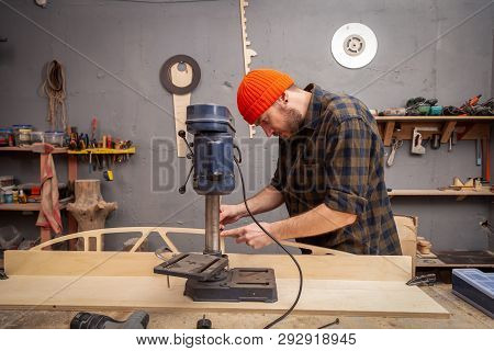 A Man Carpenter In A Hat And A Shirt  Is Carving A Wooden Board On A Large Drilling Machine In A Wor