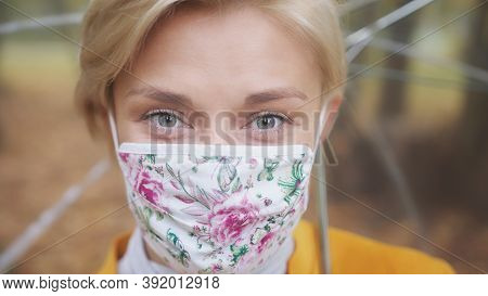 Young Blond Woman With Fashionable Face Mask And Yellow Coat Holding Transparetn Umbrella In Park. H