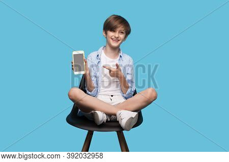Young Brunette Boy Is Sitting In An Armchair And Pointing To His Phone Screen Smiling On A Blue Stud