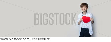 Young Boy Is Holding A Heart From Paper And Smile On A Gray Studio Wall With Free Space In Casual Cl