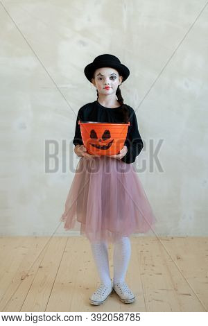 Portrait of content braided girl in tut skirt holding orange bucket for Halloween sweets against wall