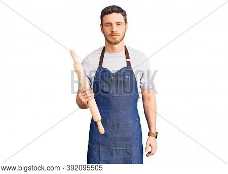 Handsome young man with bear wearing professional baker apron holding kneading roll thinking attitude and sober expression looking self confident 