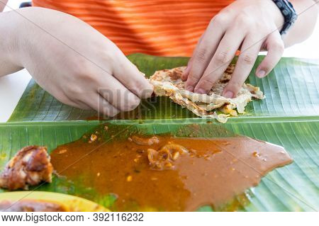 Person Enjoying Eating Roti Canai Served With Curry On Banana Leaf Using Hand