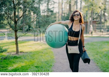 Outdoor Shot Of Happy Brunette Slim Woman Dressed In Active Wear Trendy Sunglasses Carries Sport Equ