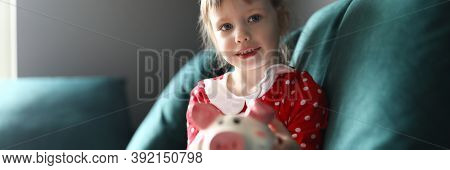 Portrait Of Smiling Beautiful Child Holding Piggy Bank Sitting On Sofa Indoors. Lovely Blonde Kid Sa
