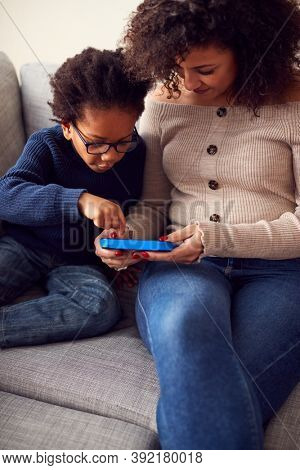 Mother And Son Sitting On Sofa At Home Playing Computer Game Together On Hand Held Device At Home