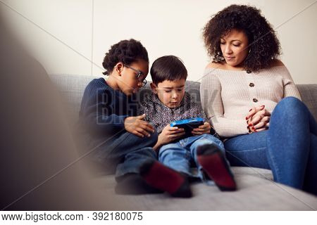 Mother And Sons Sitting On Sofa At Home Playing Computer Game Together On Hand Held Device At Home