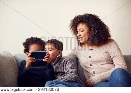 Mother And Sons Sitting On Sofa At Home Playing Computer Game Together On Hand Held Device At Home