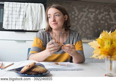 Young Woman Relaxing At Home With Antistress Coloring. She Is Sitting In The Kitchen, Eating Vookies