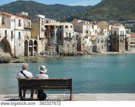 Cefalu, Sicily, Italy. May 10, 2017. A Couple Siting In The Harbot Of Cefalu. Cefalu Panoramic