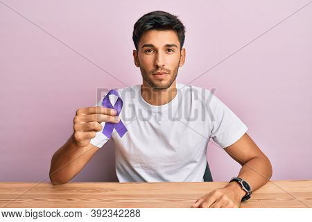 Young handsome man holding purple ribbon awareness thinking attitude and sober expression looking self confident 