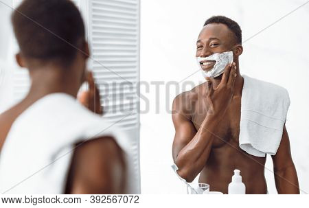 Shirtless African Guy Applying Shave Foam On Face Preparing For Shaving Standing Near Mirror In Bath