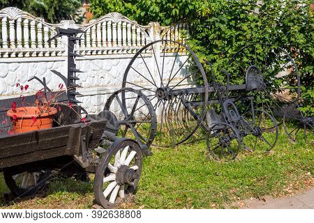Old Peasant Exhibits In Landscape Design. An Old Wooden Cart And Forged Tools For Agriculture. Russi