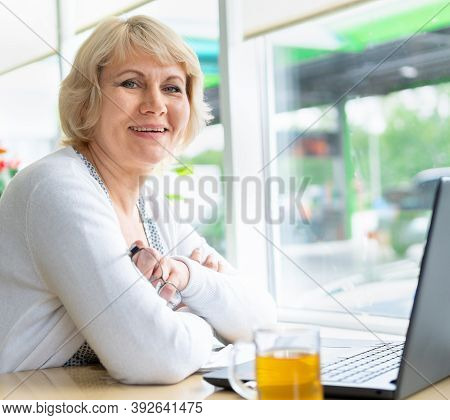 A Woman With A Laptop Is Working At A Table Near The Window.