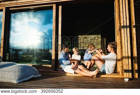 Family With Small Children Sitting On Patio Of Wooden Cabin, Holiday In Nature Concept.