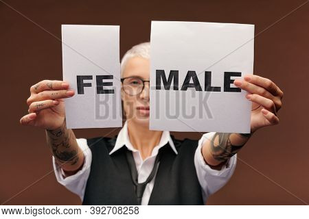 Hands of young elegant manlike woman with tattoos on arms showing two pieces of paper with syllable fe and word male in front of camera