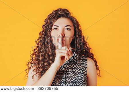 A Woman With Curly Hair Put Her Finger To Her Lips Against A Yellow Background. A Gesture Of Silence