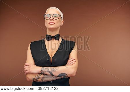 Mature blond manlike female in black waistcoat and bowtie crossing her arms covered with tattoos on chest while standing in front of camera
