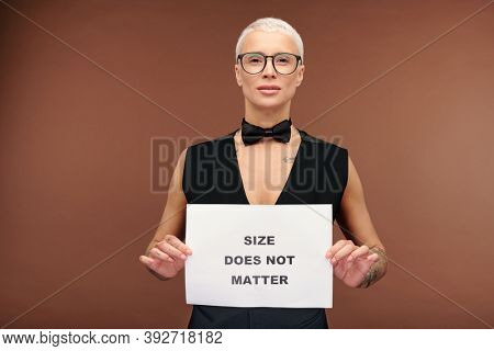 Manlike woman with short blond hair and in black bowtie and waistcoat on nude body holding paper poster saying that size does not matter