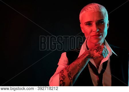 Blond elegant manlike female with tattoed arm looking at you while standing in front of camera against black background in darkness
